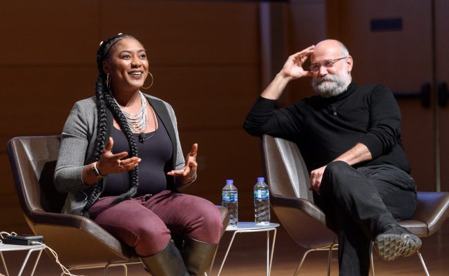 Two panelists on stage, one person is talking while the other is listening and smiling.