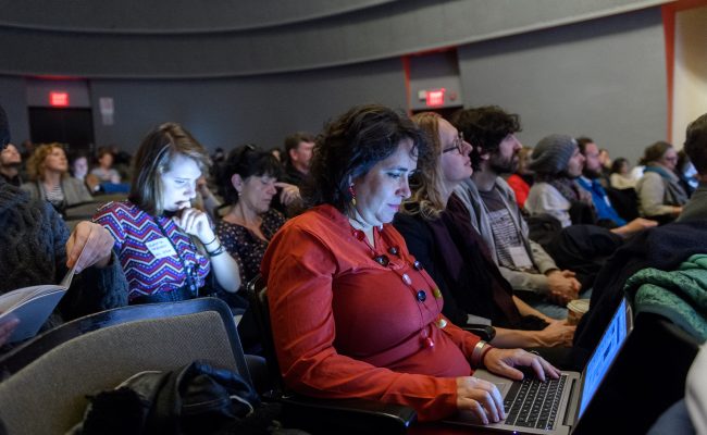 Woman sitting in an auditorium among many other people, looking down at her laptop and working.